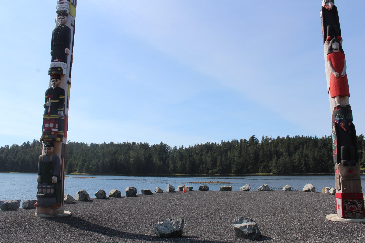 Two colorful totem poles by a river with a forest in the background and rocks on the ground.