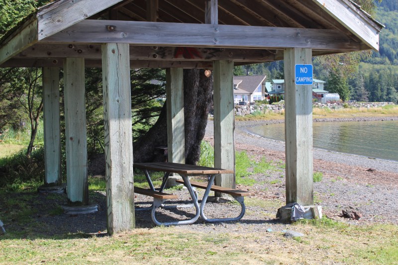 Picnic table under wooden shelter by a lake with 'No Camping' sign.