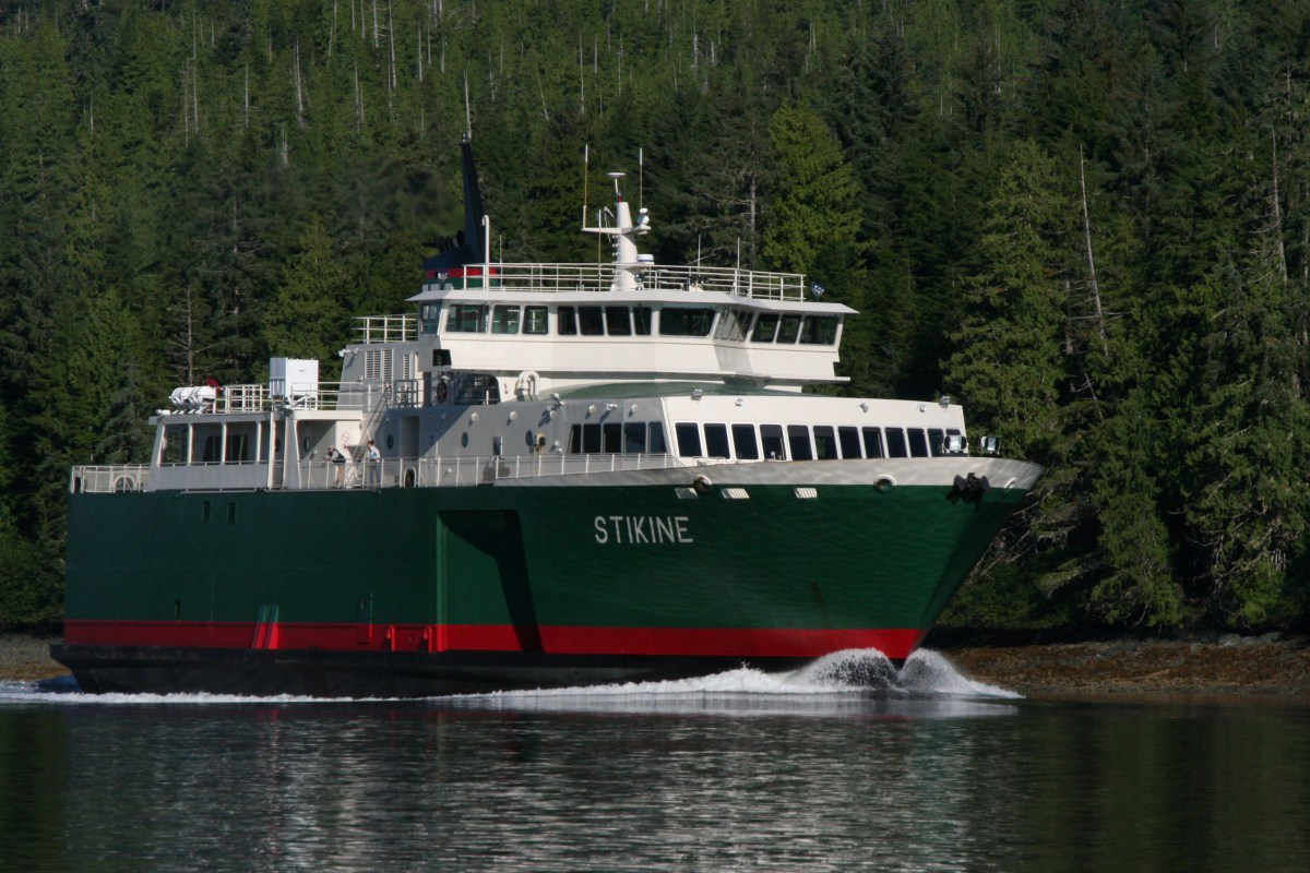 Green and white ferry named Stikine sailing near a forested shoreline.