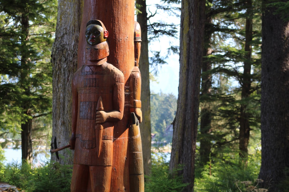 Two wooden totem poles in a forest setting with trees and greenery.