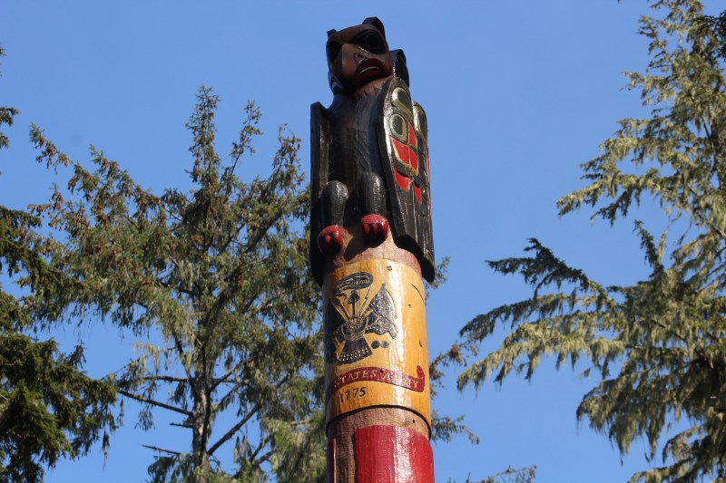 Top of a totem pole with a bear carving against a blue sky and pine trees.