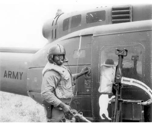 Soldier in helmet standing by military helicopter, rifle mounted beside him.